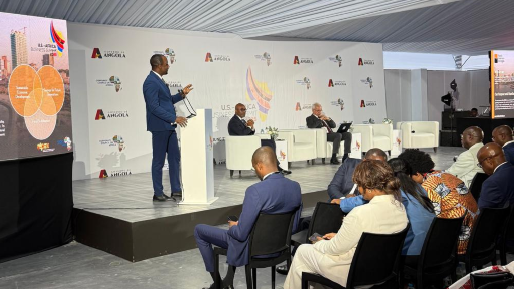 Orador em palco no U.S.–Africa Business Summit 2025 em Luanda, com painel de convidados e audiência empresarial