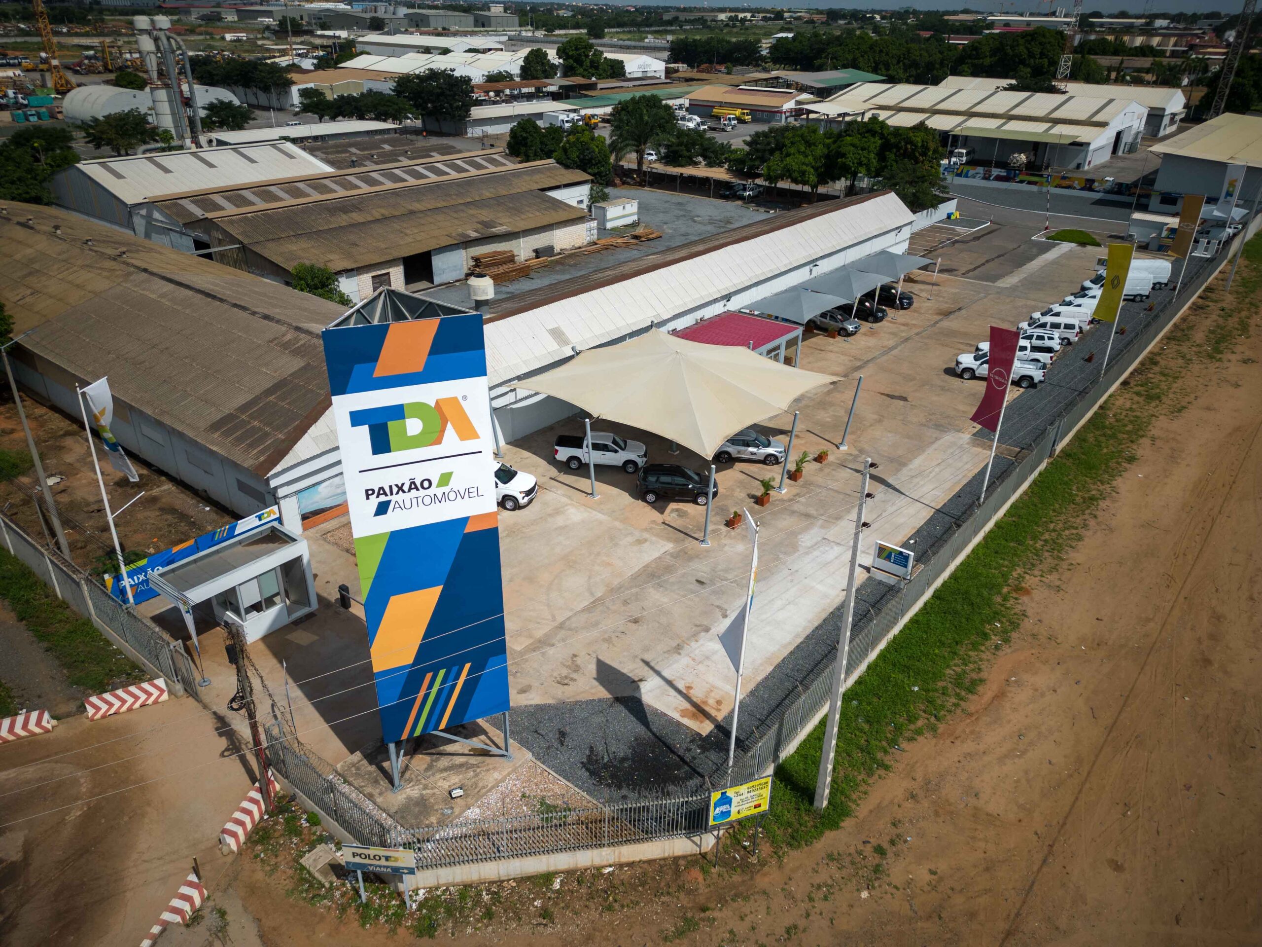 Imagem aérea do polo TDA Viana em Luanda, com área de exposição exterior, sinalética institucional e viaturas novas em parque.